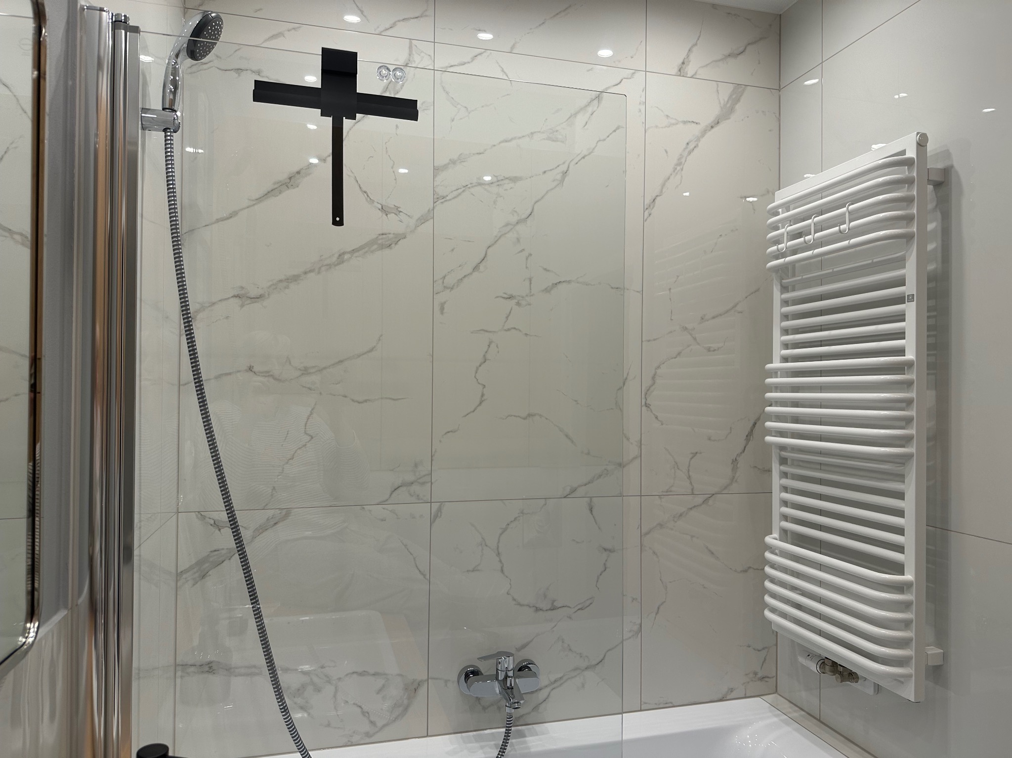 Nowoczesna łazienka z białą armaturą i płytkami imitującymi marmur, widoczny grzejnik drabinkowy i kabina prysznicowa z czarnymi detalami.