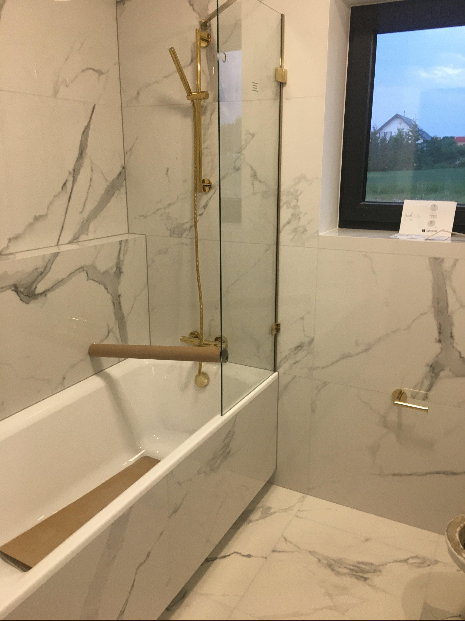 Nowoczesna łazienka z wanną obłożoną marmurowymi płytkami, złotą armaturą i szklanym panelem prysznicowym; widok przez okno na dom i zieleń.