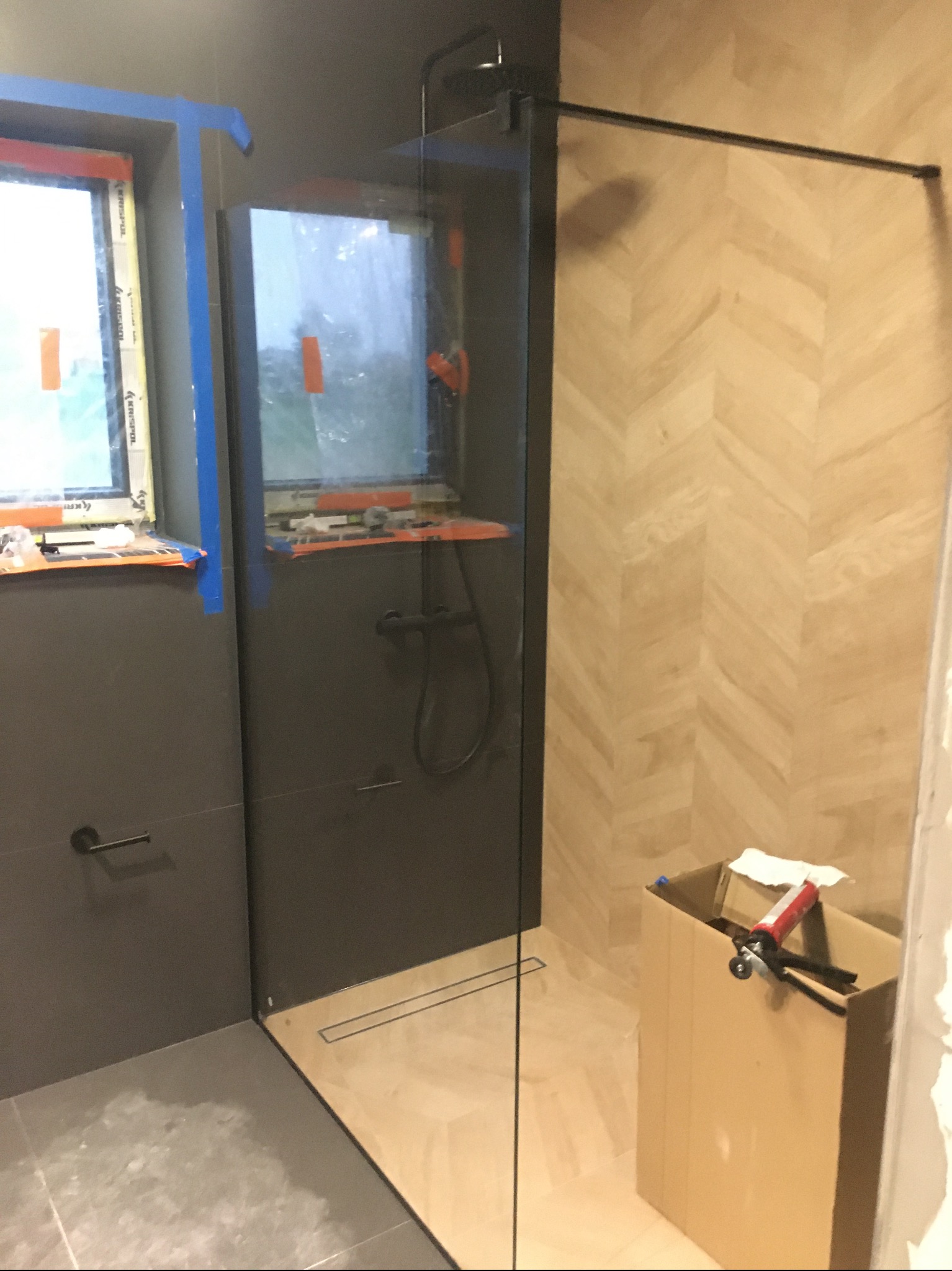 Szklana kabina prysznicowa typu walk-in z czarną armaturą, zamontowana w łazience z szarą płytką ścienną i drewnopodobną w jodełkę na ścianie prysznica. Widoczne okna z zabezpieczeniami i karton...