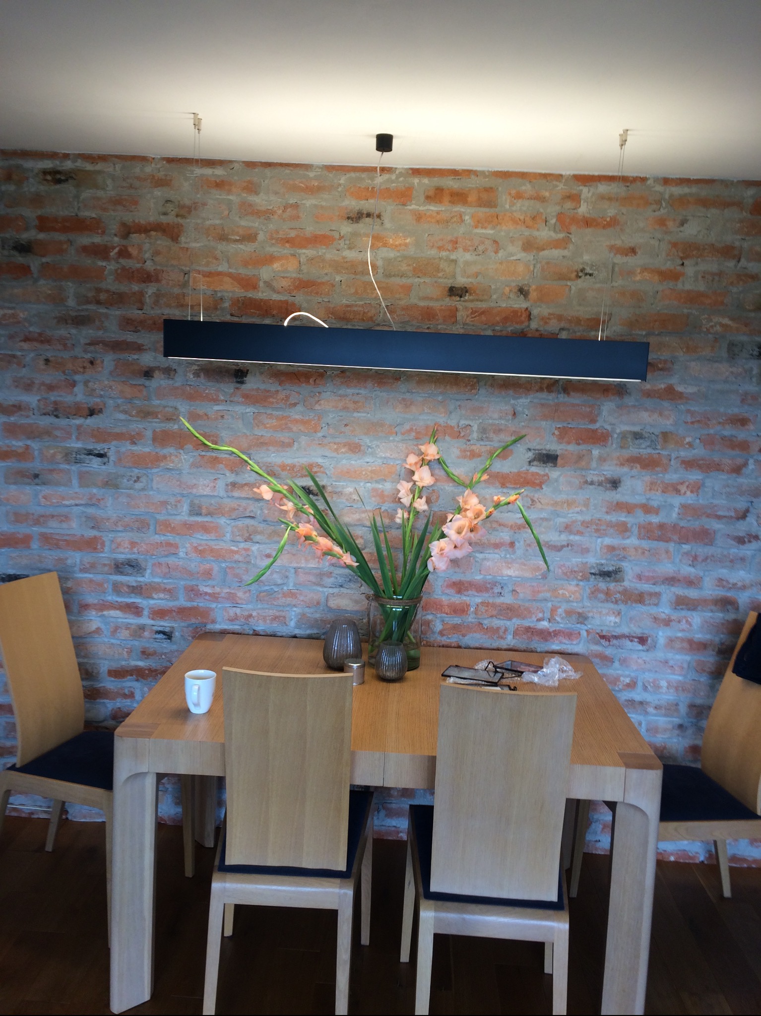 Jadalnia z drewnianym stołem, krzesłami i bukietem mieczyków na tle ściany z cegły, oświetlona podłużną lampą sufitową.