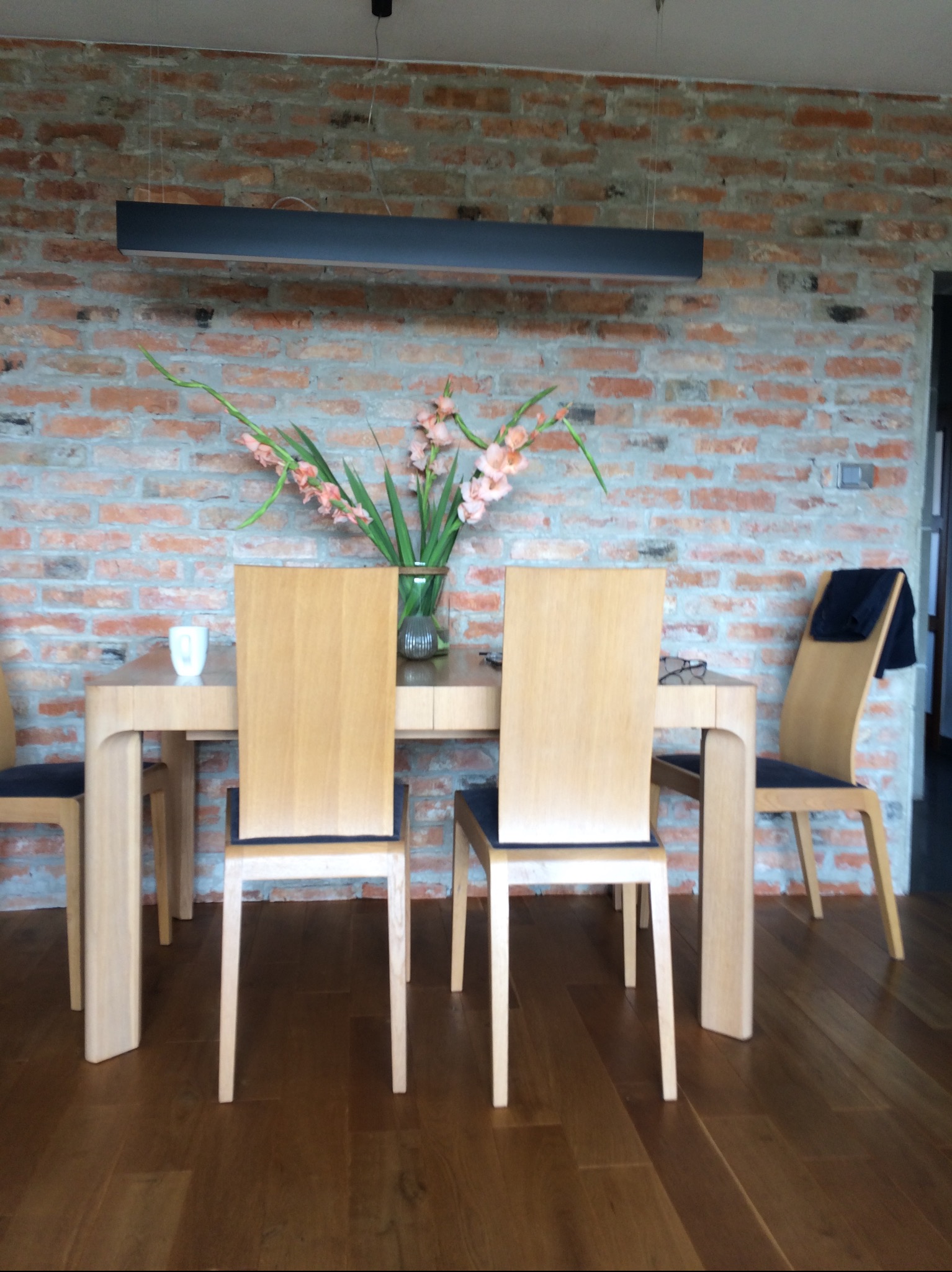 Jadalnia w stylu loft z ceglaną ścianą, drewnianym stołem i krzesłami, oświetlona podłużną lampą. Na stole wazon z kwiatami.