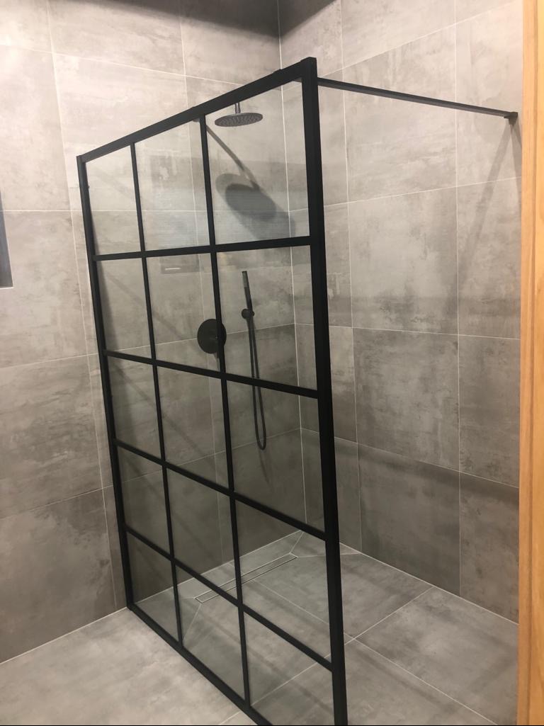 Nowoczesna kabina prysznicowa z czarnymi profilami i deszczownicą, szare płytki imitujące beton, minimalistyczny odpływ liniowy w podłodze.