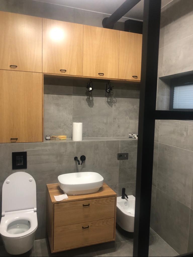 Nowoczesna łazienka z szarą płytką, drewnianymi szafkami wiszącymi i pod umywalką, białą umywalką nablatową, toaletą i bidetem, czarną armaturą i oświetleniem punktowym.