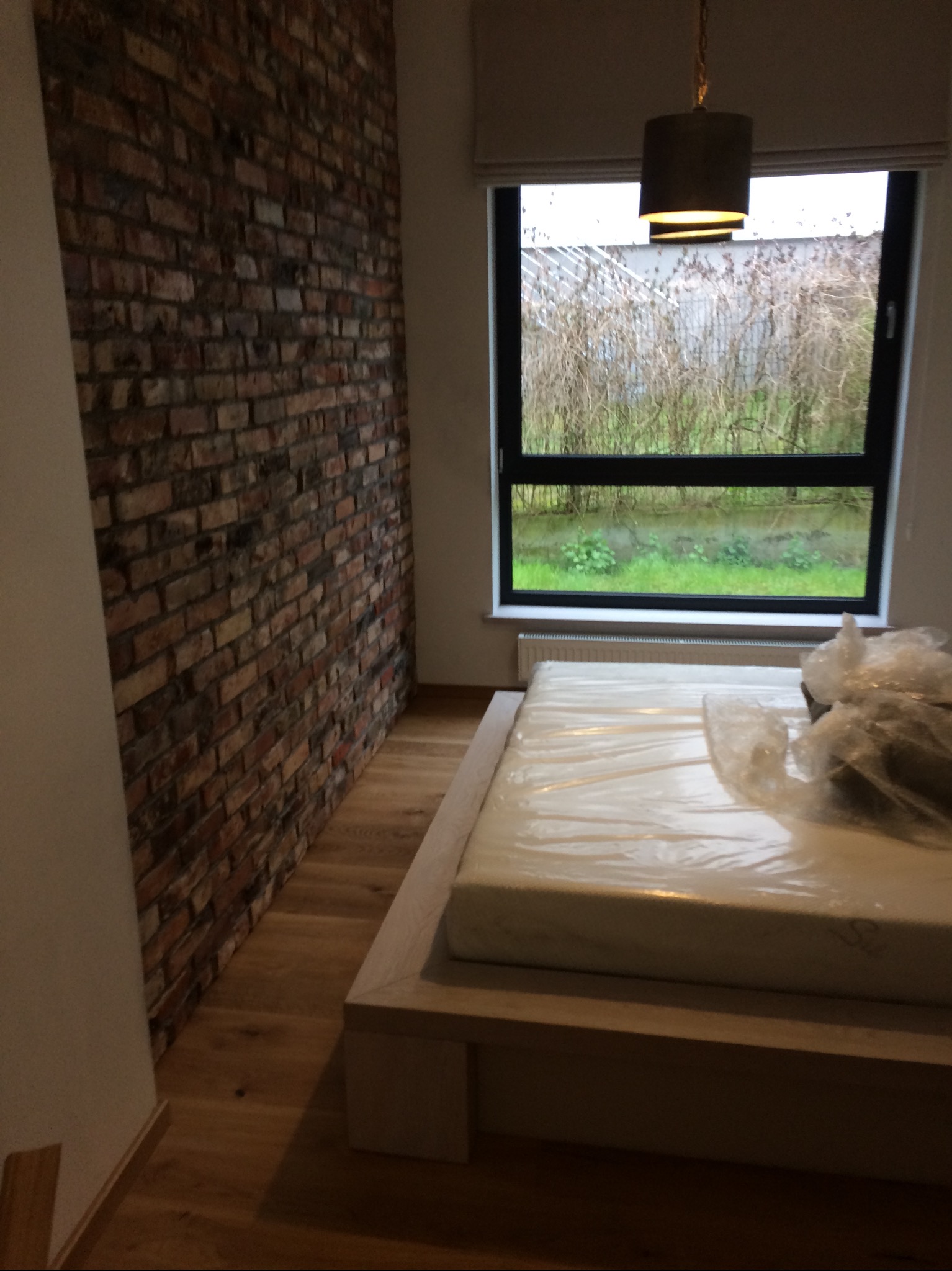 Pokój z wykończoną ścianą z cegły, drewnianą podłogą i oknem z widokiem na zieleń, z niezmontowanym łóżkiem w folii ochronnej.