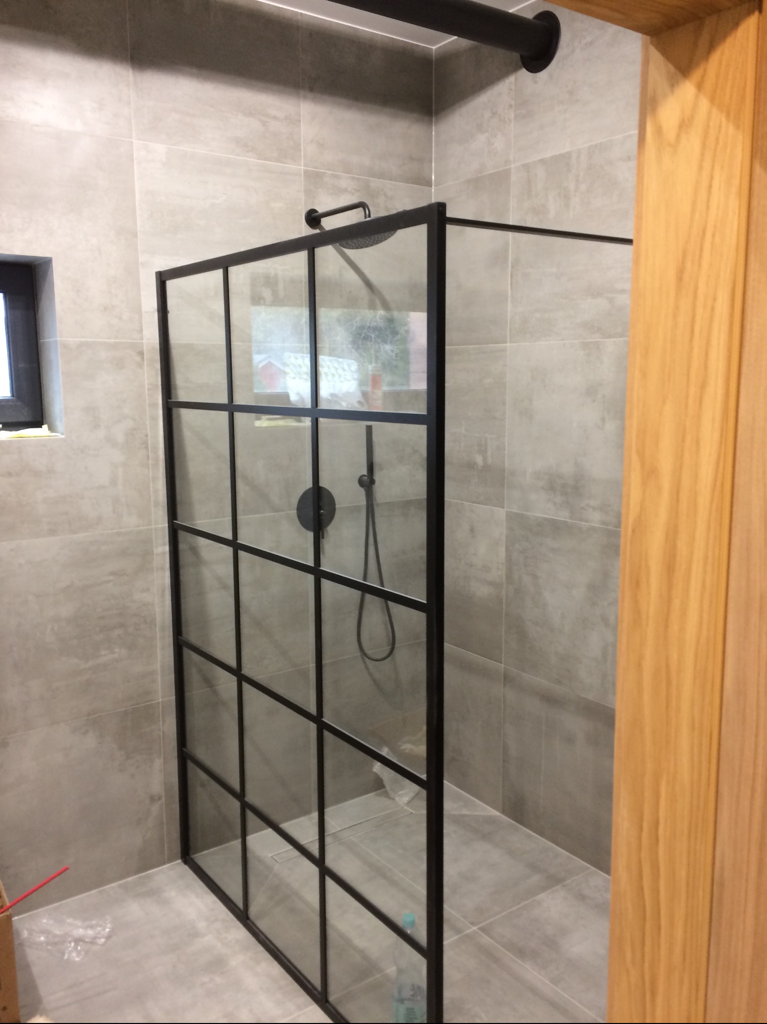 Wykończona łazienka z kabiną prysznicową typu walk-in z czarnymi profilami i szklanymi ściankami, szara płytka na ścianach i podłodze, widoczny fragment drewnianej ościeżnicy.