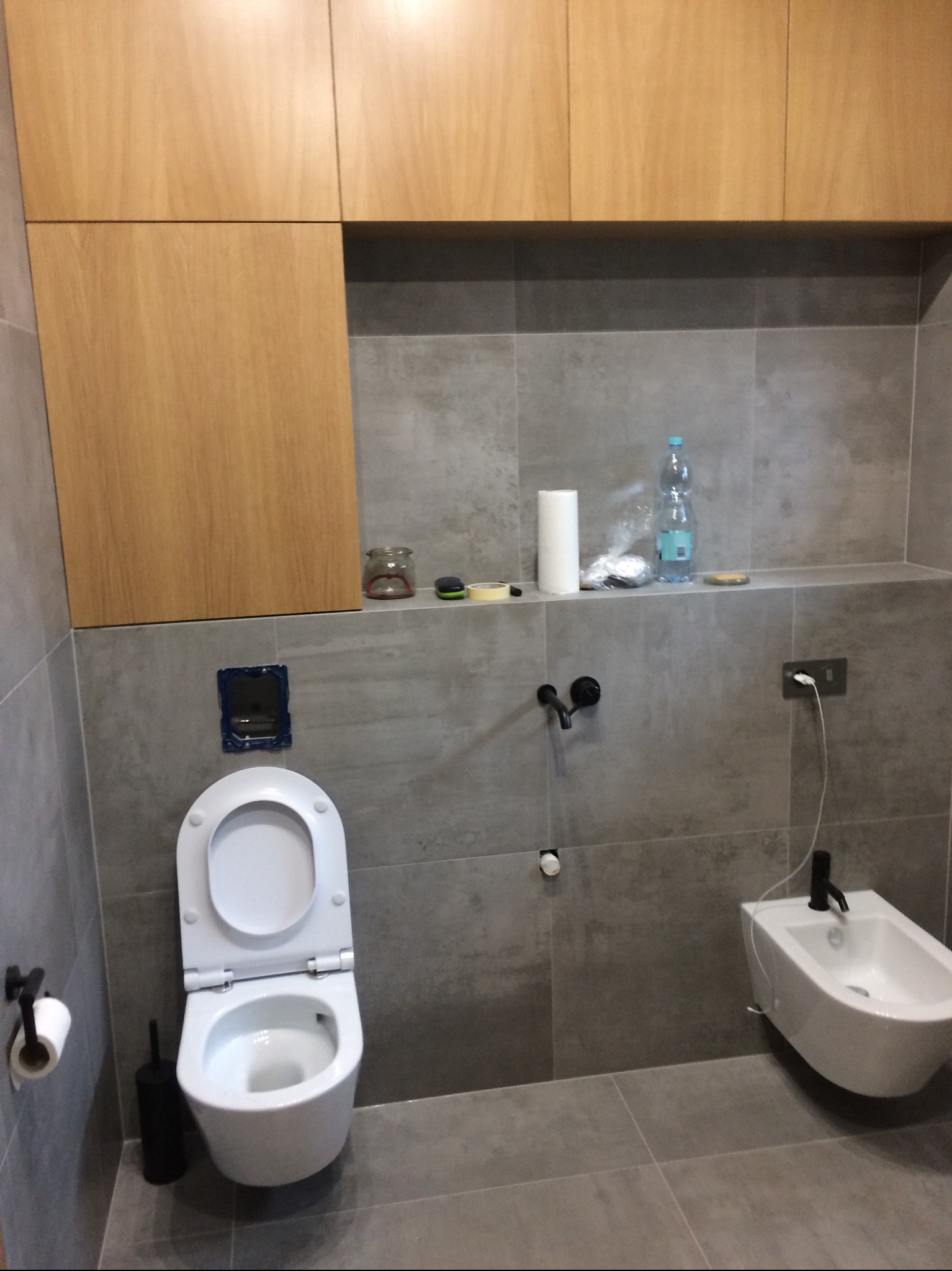 Szara łazienka z drewnianymi szafkami, toaletą i bidetem. Na półce nad toaletą akcesoria łazienkowe i butelka wody.