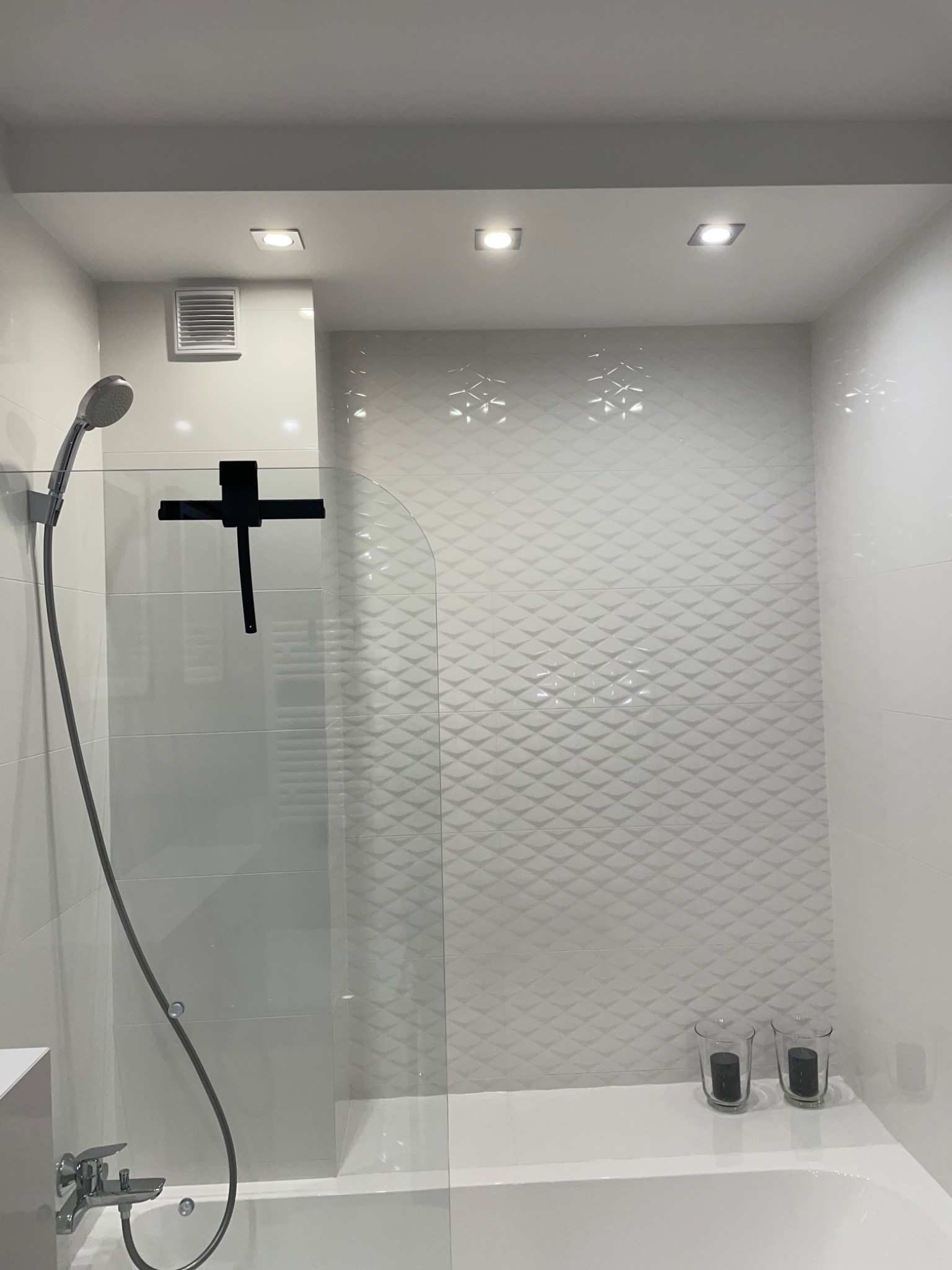 Biała łazienka z wanną, szklanym parawanem prysznicowym z czarnym uchwytem, deszczownicą i ozdobnymi płytkami ściennymi w romby, oświetlona punktowymi lampami sufitowymi.