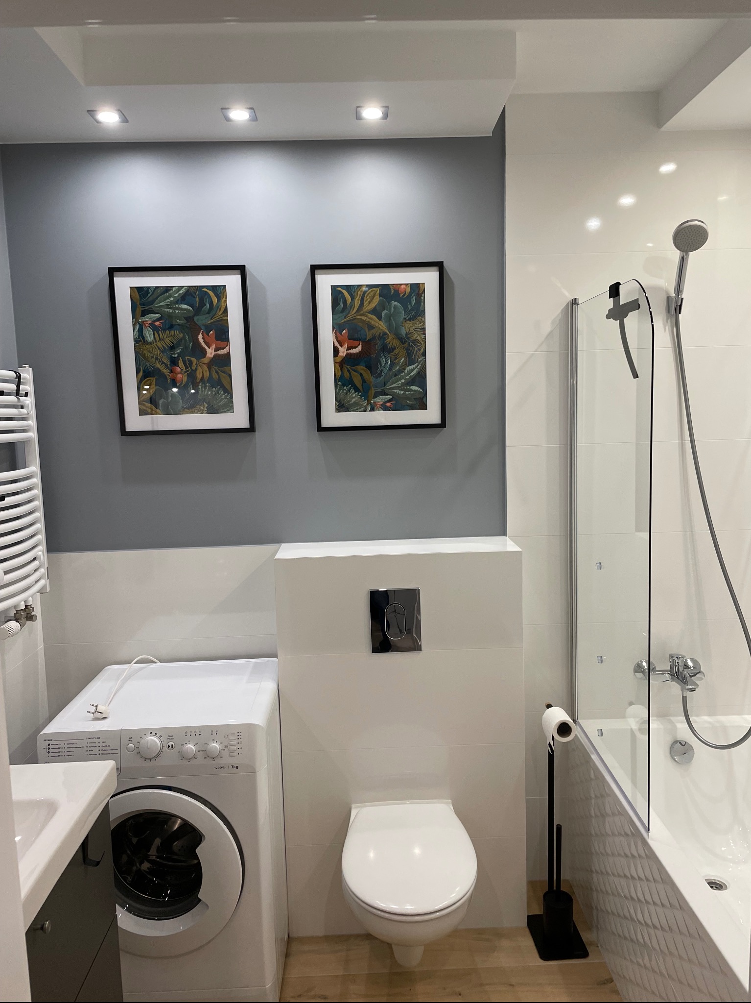 Nowoczesna łazienka z pralką obok umywalki, toaleta w zabudowie, wanna za szklanym parawanem i dwa obrazy z motywem tropikalnym na szarej ścianie.
