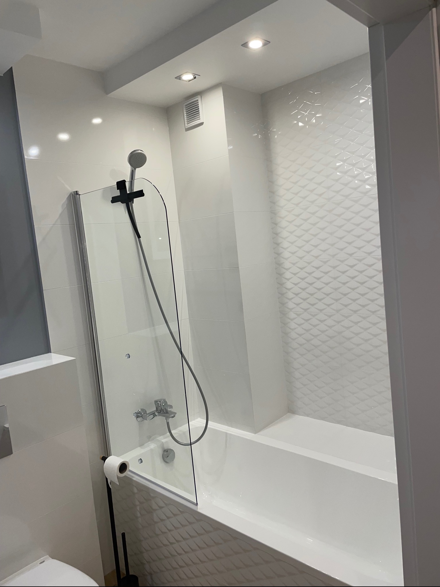 Nowoczesna łazienka z białą armaturą, szklanym parawanem nawannowym i dekoracyjnymi płytkami 3D na ścianie w strefie kąpieli.