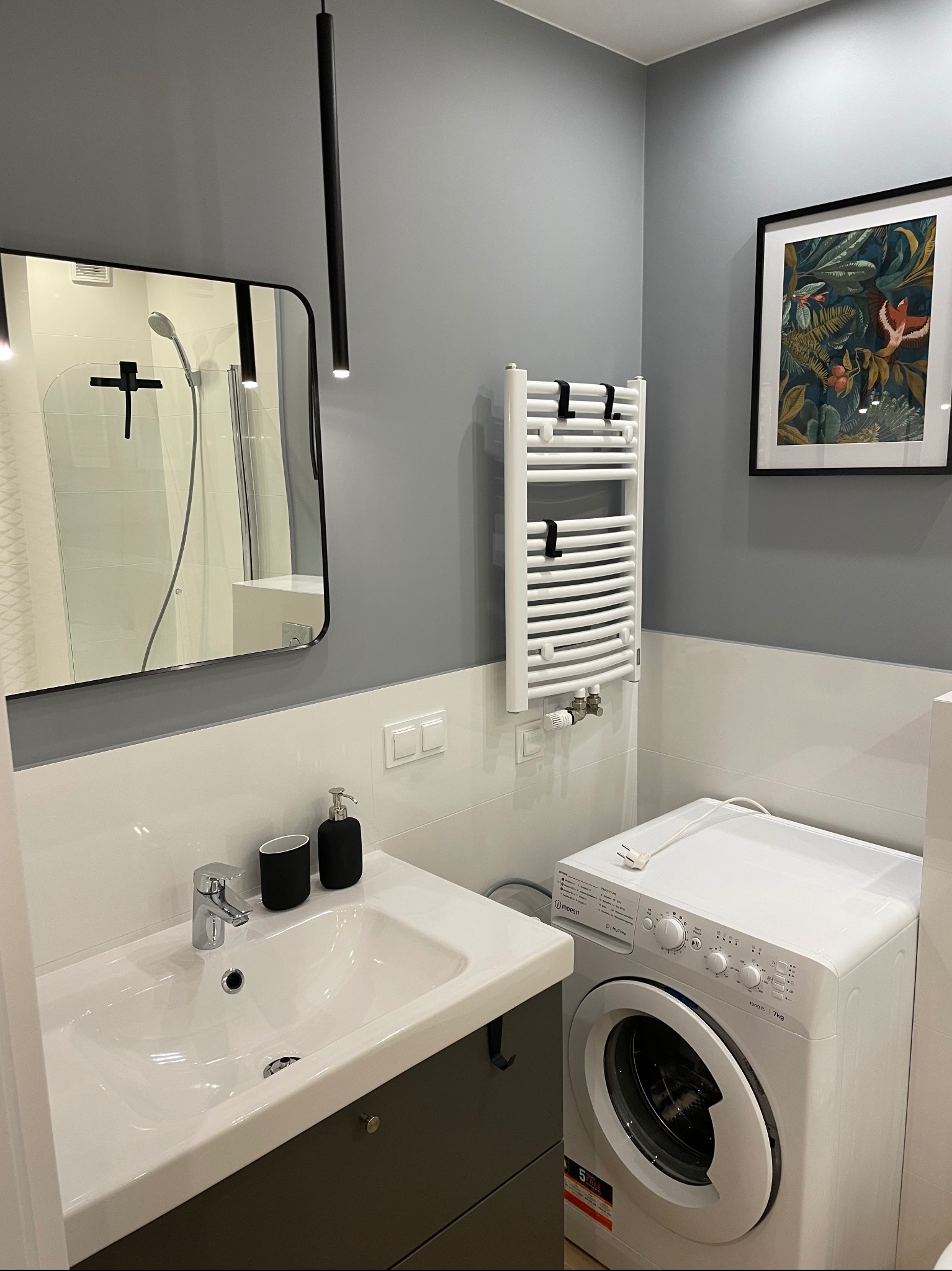 Nowoczesna łazienka w szaro-białej tonacji z umywalką nablatową, grzejnikiem drabinkowym i pralką Indesit, uzupełniona czarnymi akcentami w postaci armatury i ramy lustra.