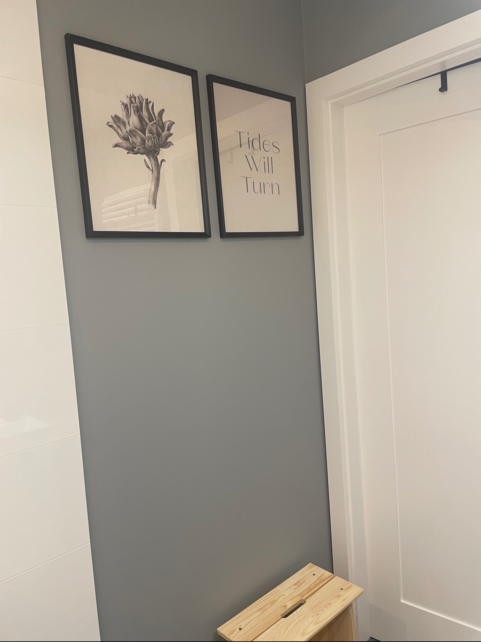 Szara ściana w łazience z dwoma czarno-białymi obrazkami w czarnych ramkach: jeden z karczochem, drugi z napisem 'Tides Will Turn', obok białe drzwi i drewniany stołek.