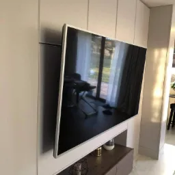 Telewizor zawieszony na ścianie z zabudową meblową w odcieniach bieli i brązu, widoczne elementy dekoracyjne i odbicie okna.