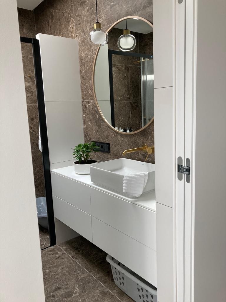 Nowoczesna łazienka z białymi szafkami pod umywalką, złotą armaturą, okrągłym lustrem i marmurowymi płytkami na ścianie, oświetlona wiszącymi lampami ze szklanymi kloszami.