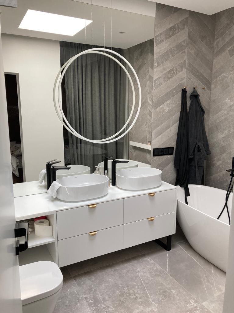 Nowoczesna łazienka z podwójną umywalką nablatową na białej szafce z złotymi uchwytami, okrągła lampa wisząca, szara ściana w jodełkę i wolnostojąca wanna.