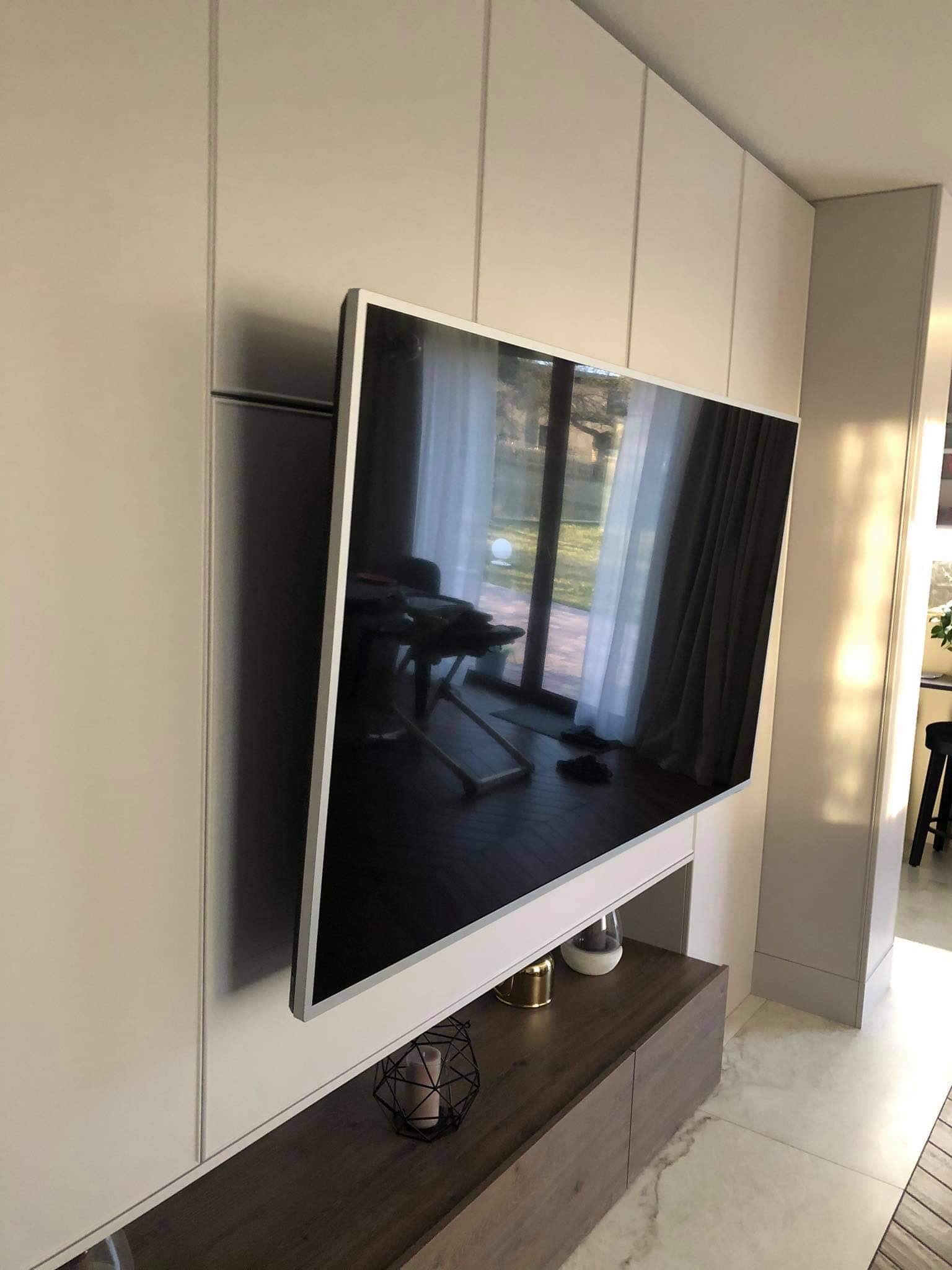 Telewizor zawieszony na ścianie z zabudową meblową w odcieniach bieli i brązu, widoczne elementy dekoracyjne i odbicie okna.