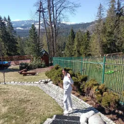 Dziewczyna w dresie macha ręką na tle zadbanego ogrodu z kamienną ścieżką, zielonym płotem i widokiem na góry w oddali.