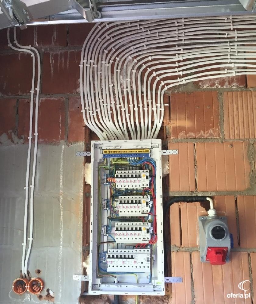 Rozdzielnica elektryczna z licznymi przewodami w białych peszlach, zamontowana na surowej ścianie z cegły, widoczna puszka instalacyjna z czerwonym gniazdem.