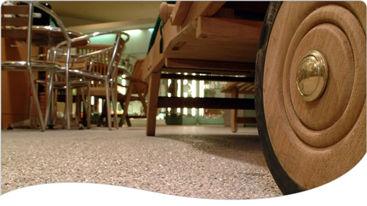 Jasna, drobnoziarnista posadzka z żywicy epoksydowej w salonie meblowym, z widocznym drewnianym wózkiem z kołem z bliska i meblami w tle.