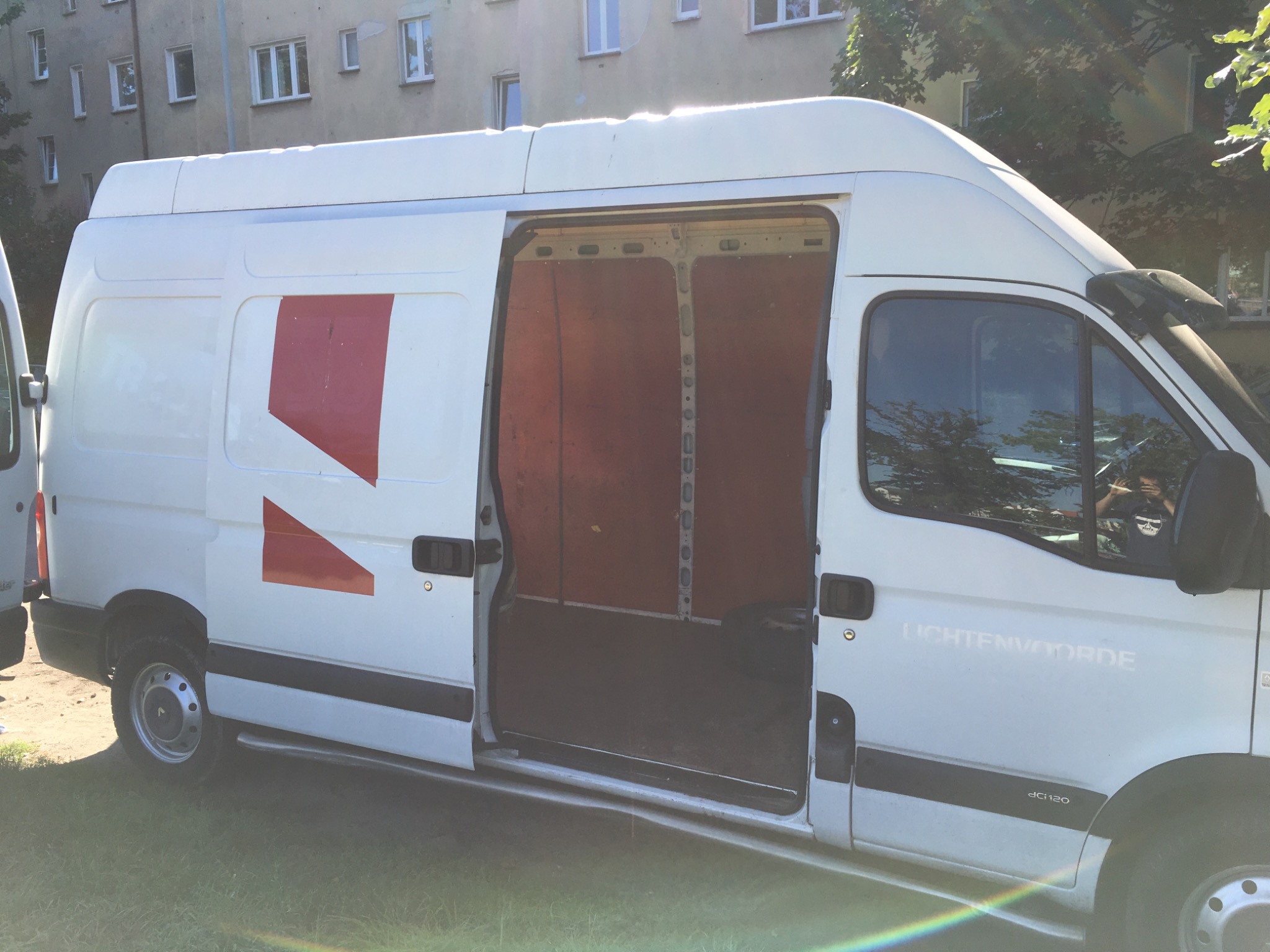 Biały bus dostawczy z otwartymi drzwiami bocznymi, widoczne puste wnętrze przestrzeni ładunkowej, na karoserii czerwone oznaczenia firmy, budynek mieszkalny w tle.