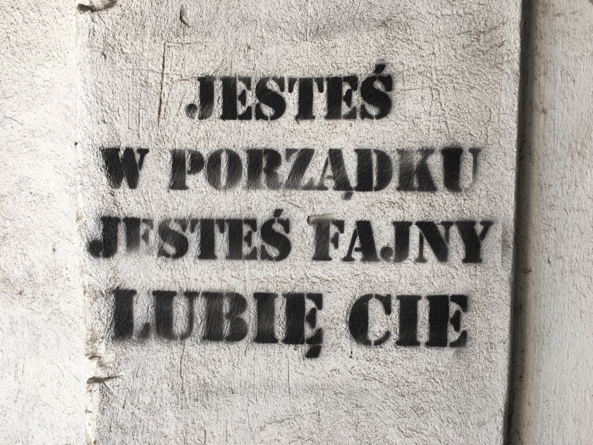 Tekst 'Jesteś w porządku, jesteś fajny, lubię Cię' namalowany czarną farbą przez szablon na szarej, chropowatej ścianie.