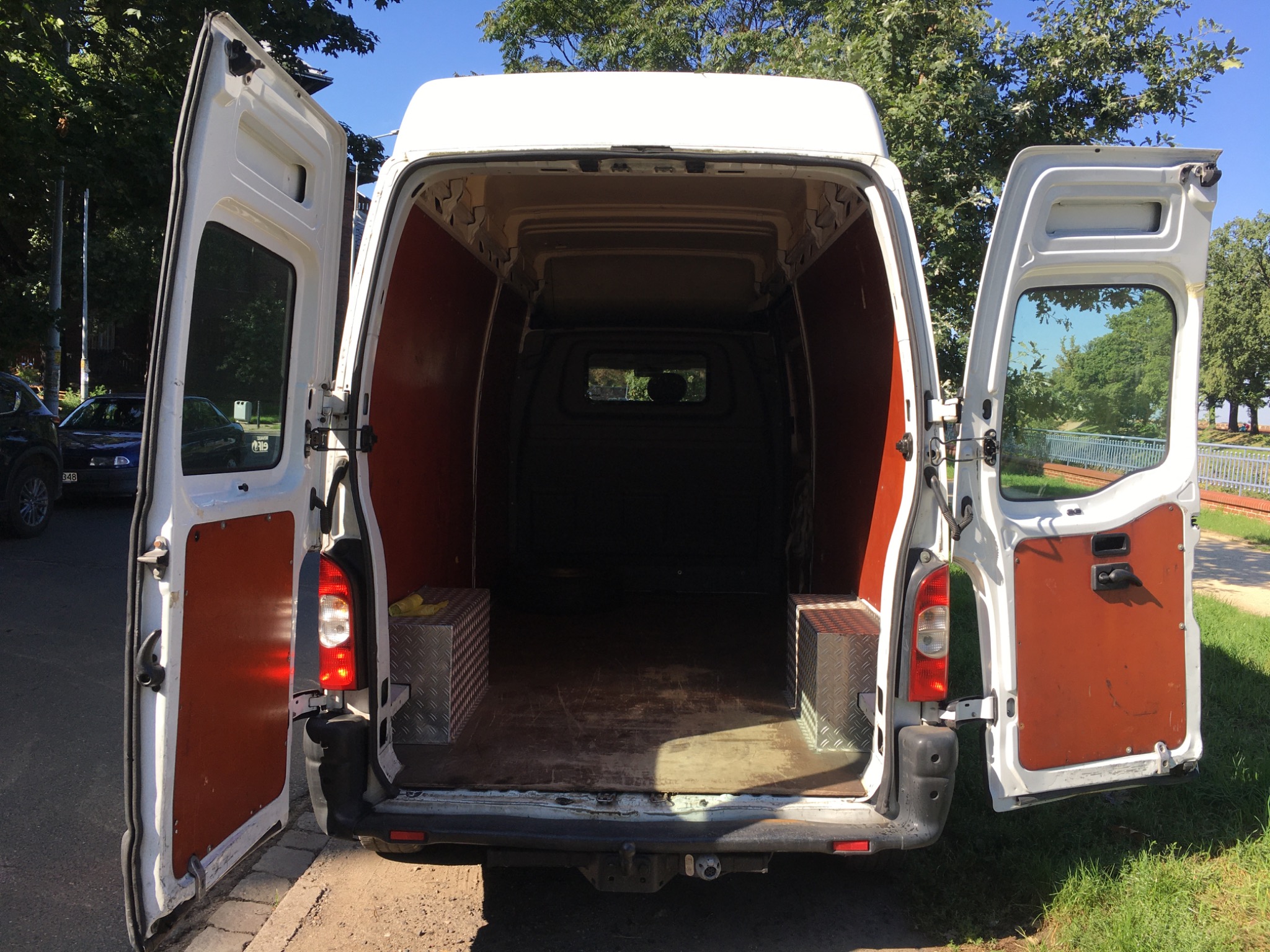 Wnętrze białego furgonetki z otwartymi tylnymi drzwiami, widoczne dwie metalowe skrzynki narzędziowe na podłodze, tło z zielenią drzew.