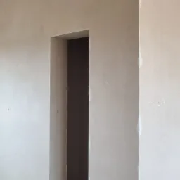 Świeżo nałożone tynki w jasnym odcieniu beżu, widoczne przewody instalacji elektrycznej i hydraulicznej przy podłodze, surowy stan wykończenia wnętrza z widocznym otworem drzwiowym.