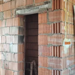 Wnętrze budynku w trakcie budowy, widoczne ściany z czerwonej cegły, instalacja elektryczna w ścianie, drabina w przejściu, oraz rury na podłodze.