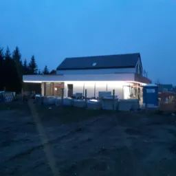 Nowoczesny dom jednorodzinny w trakcie budowy oświetlony listwami LED na zewnątrz, widoczne materiały budowlane i przenośna toaleta, sfotografowany o zmierzchu.