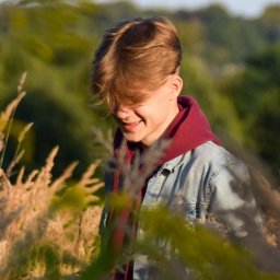 toporek_at_lens - Młody mężczyzna w jeansowej kurtce i bordowej bluzie z kapturem, uśmiechnięty, stojący w wysokiej trawie na tle zielonych drzew.