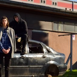 toporek_at_lens - Dwaj nastolatkowie pozują na tle rozbitego, czarnego samochodu bez kół, stojącego przed budynkiem z żółtym karmnikiem dla ptaków na ścianie.