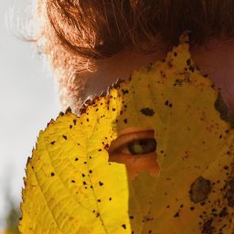 toporek_at_lens - Zielone oko kobiety spogląda przez dziurę w żółtym liściu z czarnymi plamkami, widoczne rude włosy i fragment skóry.