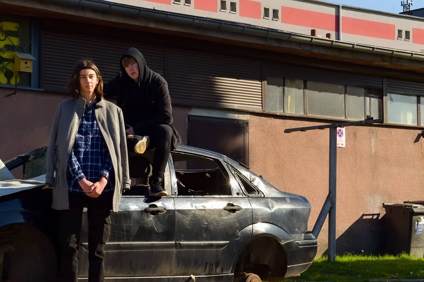 Dwaj nastolatkowie pozują na tle rozbitego, czarnego samochodu bez kół, stojącego przed budynkiem z żółtym karmnikiem dla ptaków na ścianie.