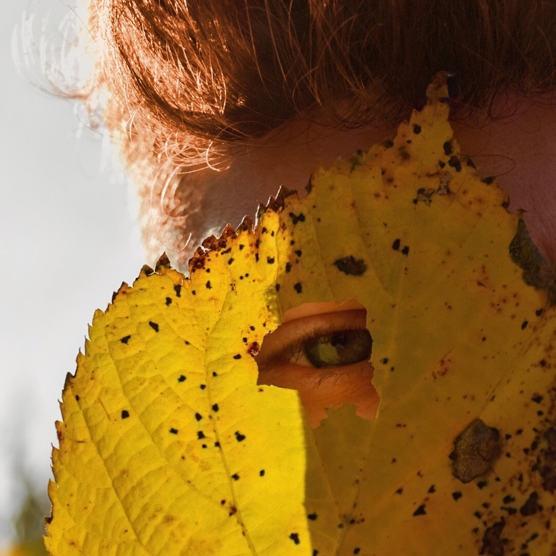 Zielone oko kobiety spogląda przez dziurę w żółtym liściu z czarnymi plamkami, widoczne rude włosy i fragment skóry.