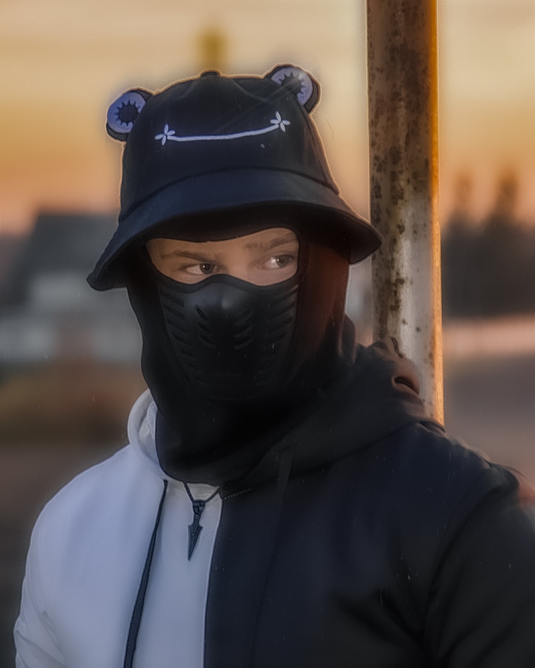 Portret osoby w czarno-białej bluzie z kapturem, czarnym kapeluszu z uszkami i maską na twarzy, stojącej obok metalowego słupka na rozmytym tle o zachodzie słońca.