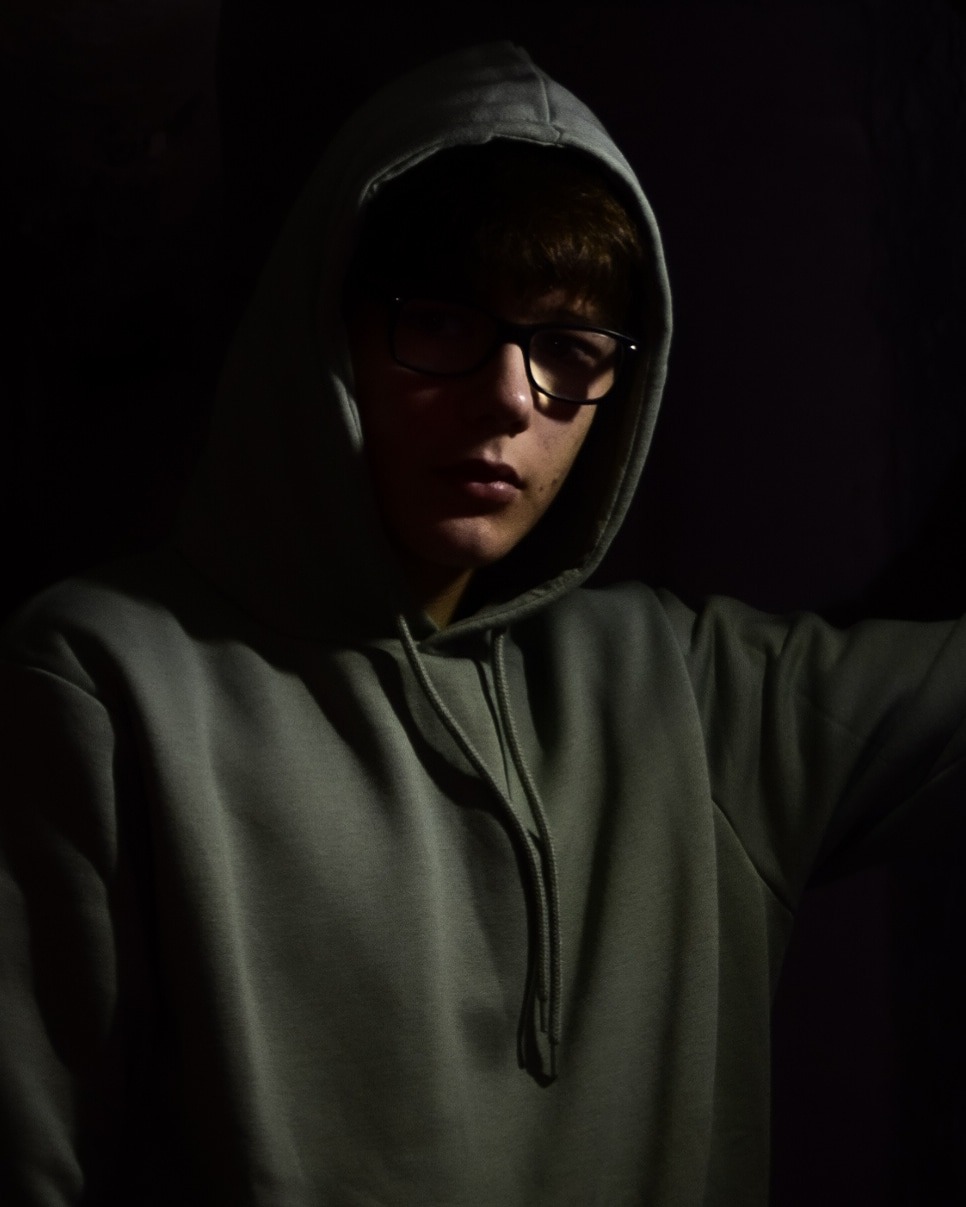 Portret młodego mężczyzny w okularach i szarej bluzie z kapturem, oświetlonego z góry, na ciemnym tle.