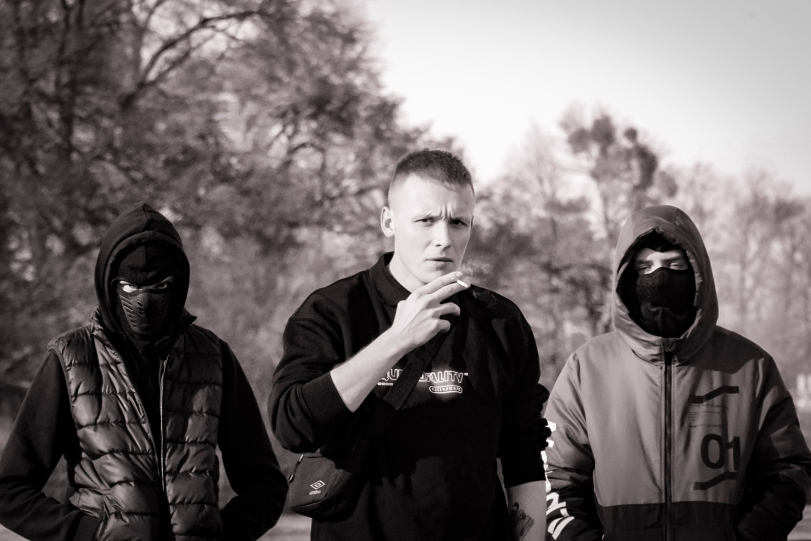 Czarno-białe zdjęcie trzech mężczyzn na zewnątrz: dwóch w maskach i kapturach, jeden z papierosem w ustach, patrzący prosto w obiektyw, tło z drzewami.