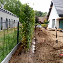 Przygotowanie terenu pod nowy trawnik z rolki: wykopany rów wzdłuż żywopłotu i ogrodzenia panelowego, widoczne narzędzia ogrodnicze i pracownik w tle.