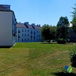 Rozległy trawnik miejski z widocznymi śladami po koszeniu, otoczony budynkami mieszkalnymi i drzewami, w słoneczny dzień.