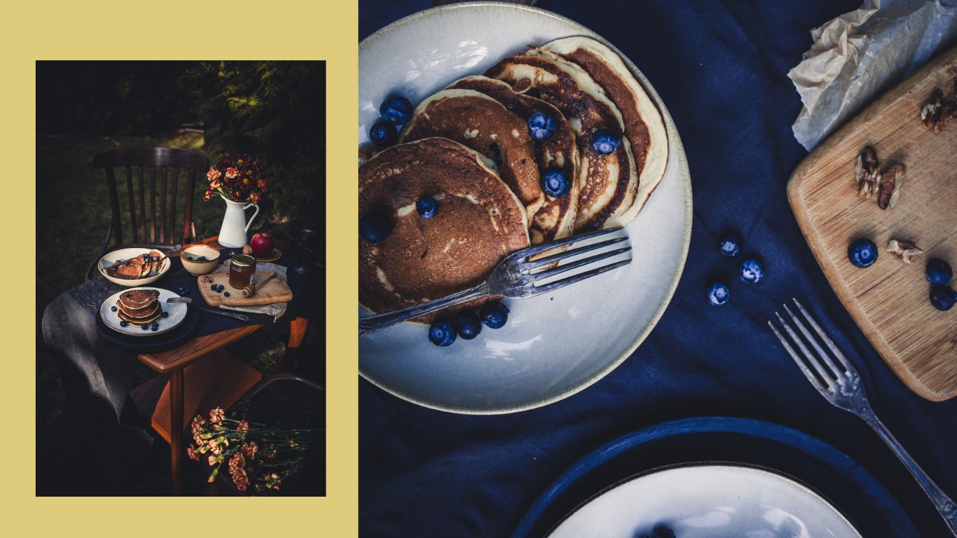 Stylizowany stół z naleśnikami i borówkami w plenerze, rustykalne naczynia, drewniana deska z orzechami, kwiaty w wazonie i słoik miodu, ciemne tło z zielenią.