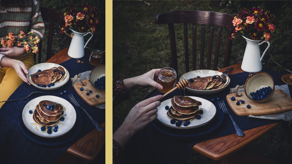 Dwa talerze z ułożonymi w stos naleśnikami, udekorowane świeżymi borówkami, na stole z ciemnym obrusem, obok dzbanek z kwiatami i słoik miodu, osoba polewa naleśniki miodem.