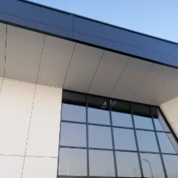 Nowoczesna fasada budynku z dużymi oknami i panelami w odcieniach szarości i bieli, widziana z dołu pod kątem, z widocznym fragmentem niebieskiego nieba.