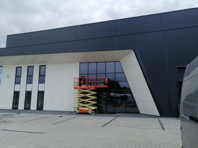 Pracownicy na podnośniku koszowym, myjący okna na wysokości biurowca w Siedlcach. Widoczna elewacja budynku, szare niebo i fragment zaparkowanego busa.