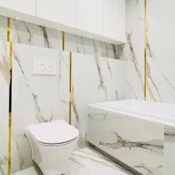 Nowoczesna łazienka wykończona białymi płytkami marmuropodobnymi z złotymi akcentami, podwieszana toaleta, zabudowa wanny i szafki wiszące z ryflowanymi frontami.