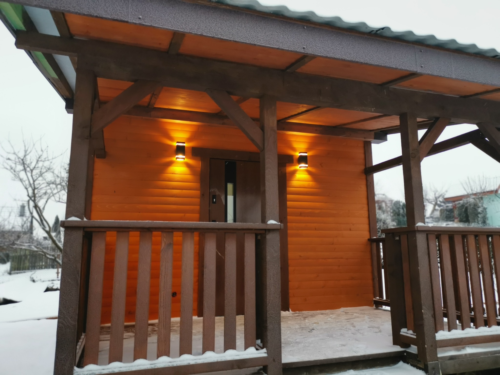 Drewniany domek z oświetleniem zewnętrznym i balustradą, pokryty śniegiem, widok z zewnątrz w pochmurny dzień.