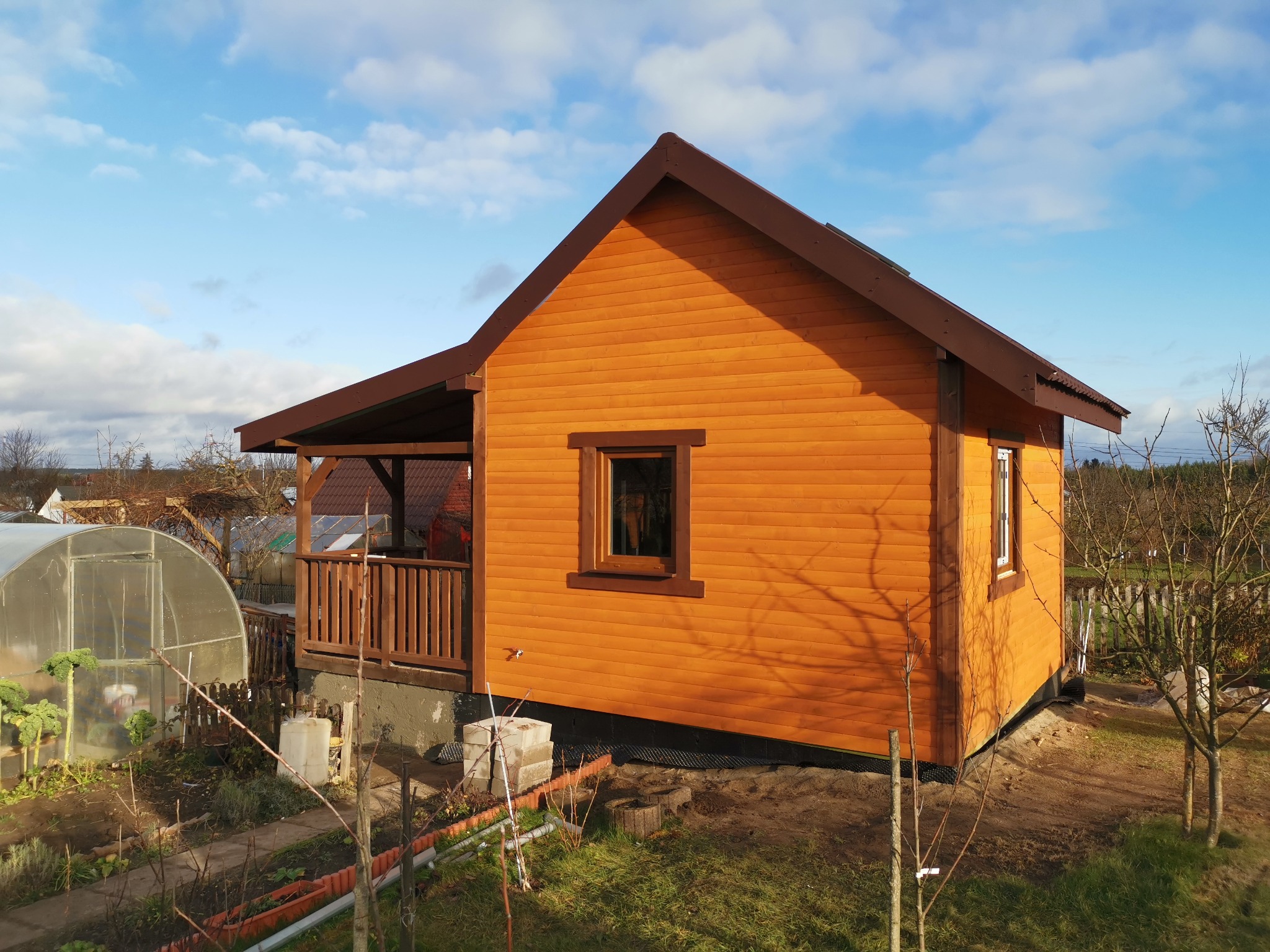 Nowo wybudowany, pomarańczowy domek letniskowy z brązowym dachem, widoczny z zewnątrz, z ogrodem w tle i szklarnią po lewej stronie.