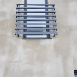 Szara, pozioma balustrada okienna z metalowych profili na tle surowej, jasnej elewacji budynku z widocznym oknem.