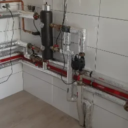 Złożona instalacja hydrauliczna z rurami miedzianymi i czerwonymi rurami plastikowymi, izolowanymi folią aluminiową, zamontowana w pomieszczeniu z białymi, kwadratowymi płytkami ściennymi i podłogą...