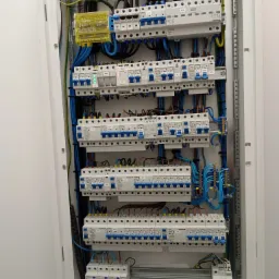 Rozdzielnica elektryczna z widocznymi zabezpieczeniami, przewodami w kolorach niebieskim, żółto-zielonym i brązowym, zamontowana w białej obudowie, z dokumentacją techniczną na dole.