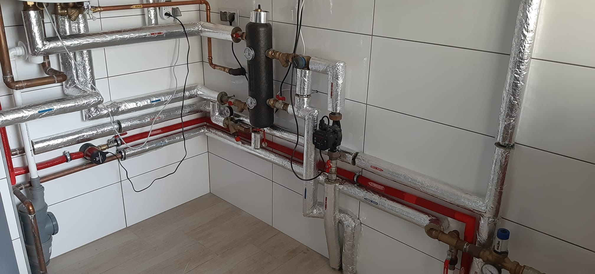 Złożona instalacja hydrauliczna z rurami miedzianymi i czerwonymi rurami plastikowymi, izolowanymi folią aluminiową, zamontowana w pomieszczeniu z białymi, kwadratowymi płytkami ściennymi i podłogą...