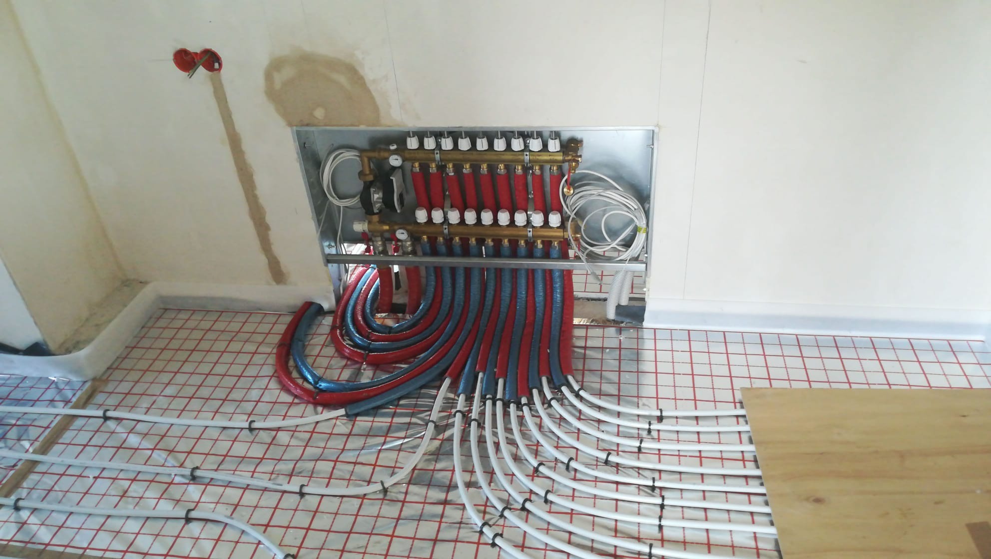 Rozdzielacz ogrzewania podłogowego z czerwono-niebieskimi rurami w trakcie montażu, widoczne białe rury na podłodze z siatką, ściana w stanie surowym.